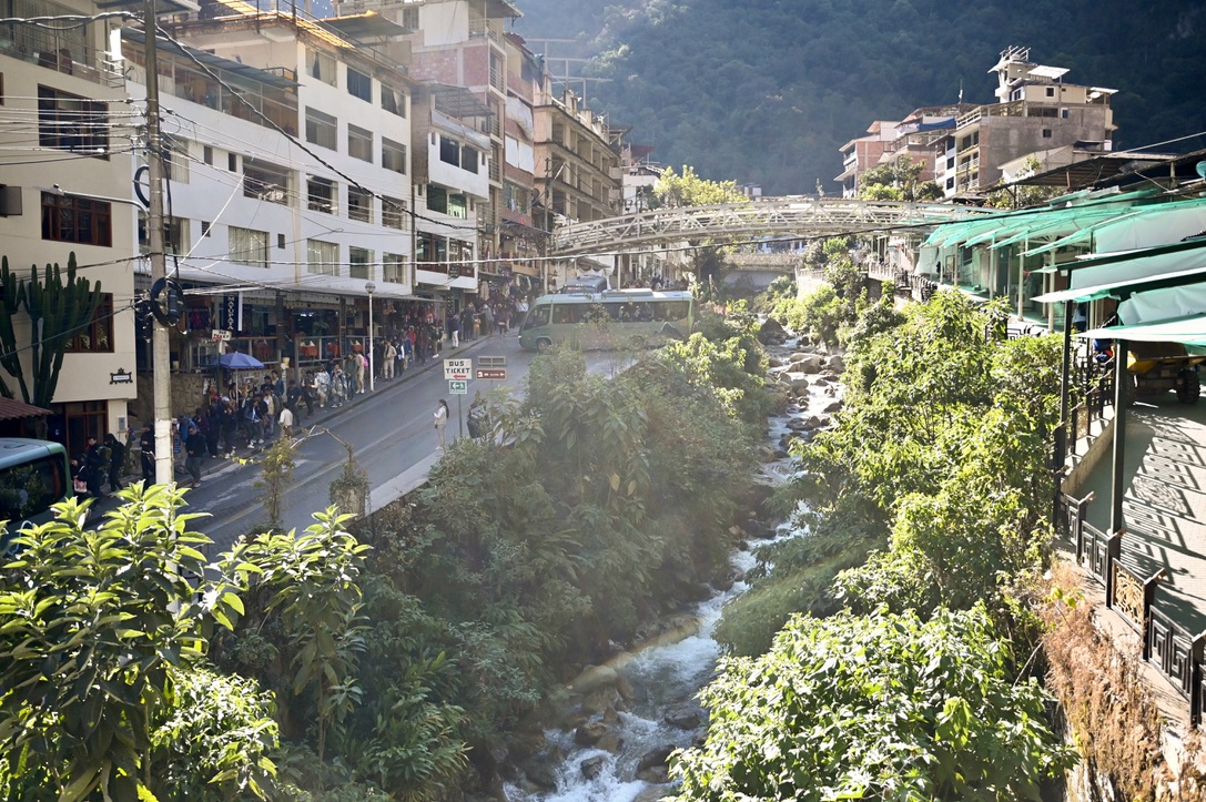 Mountain stream surrounded by lush vegetation running through a town with building stacked close together, sidewalks by commercial buildings