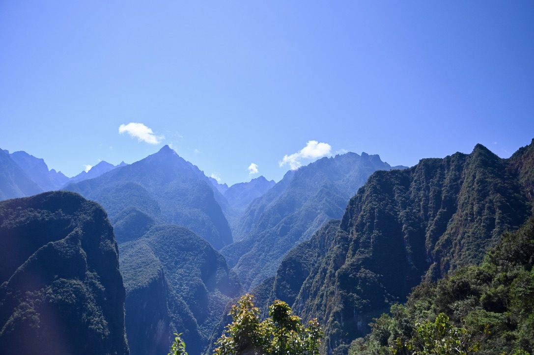 Series of sharp mountains with deep valleys covered in vegetation with hazy blue tint towards the back, clear blue sky with few clouds