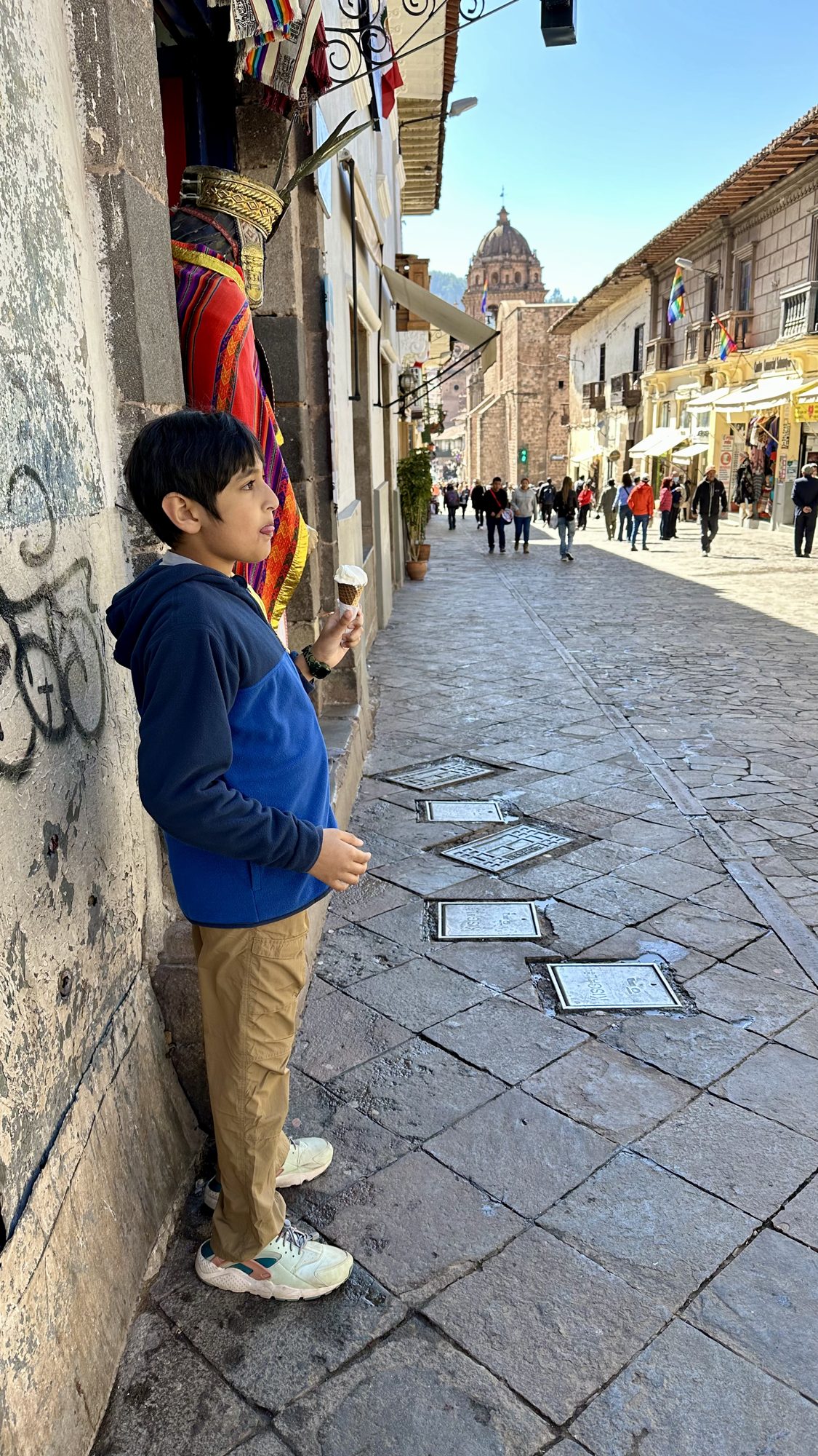 Boy standing by a shop on a sunny Cusco street, with a church tower beyond