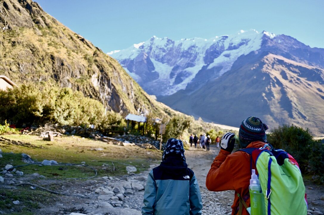 Two hikers on a rocky path toward snow-covered peaks