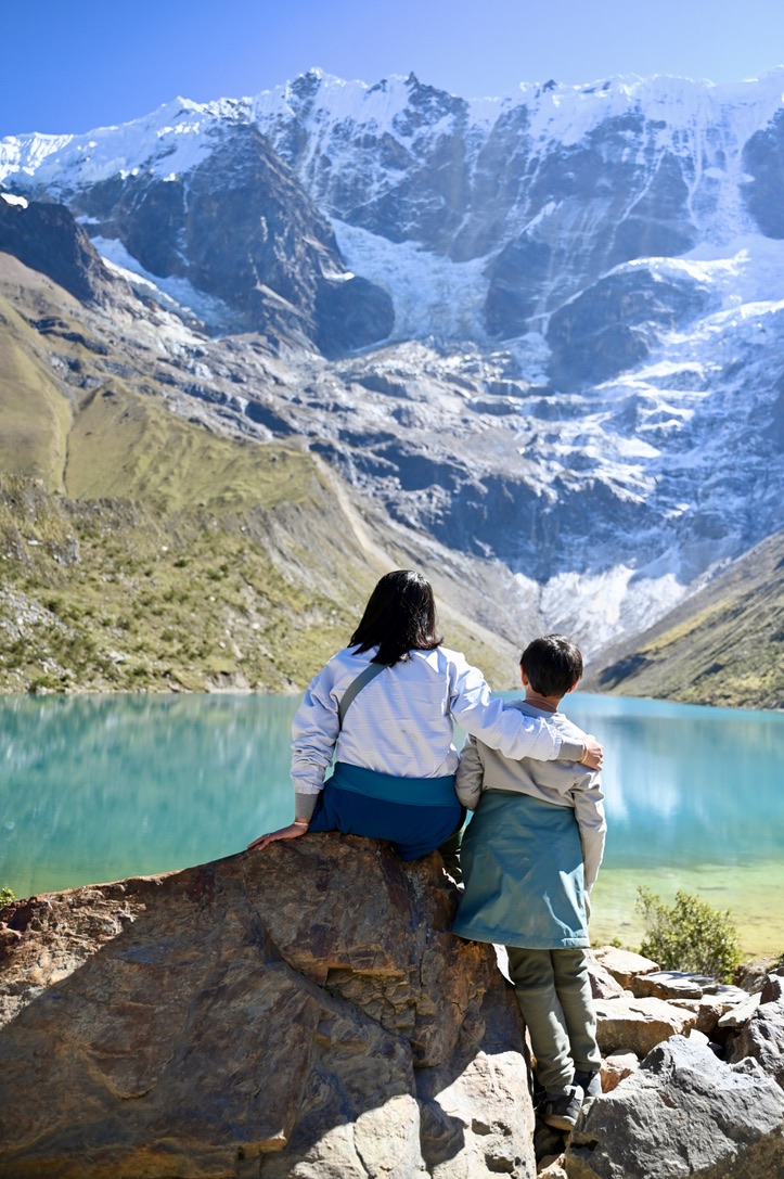 Woman and boy sitting on a rock, looking at glacier covered mountain walls, with a blue lake in the mid-ground