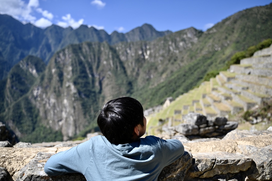 Boy with hands on a short railing wall looking at tropical mountain peaks and stone wall terraces on one of the mountain sides