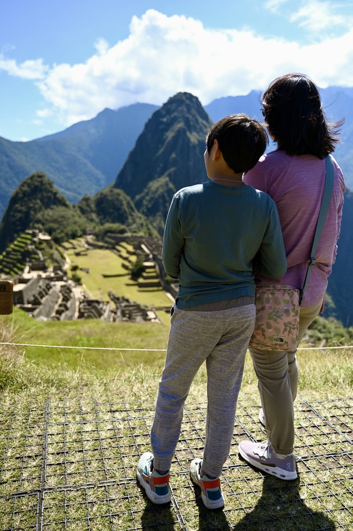 Woman and boy standing on grass and looking at ancient ruins of Machu Picchu over lush grass covered mountain top with other mountain peaks all around