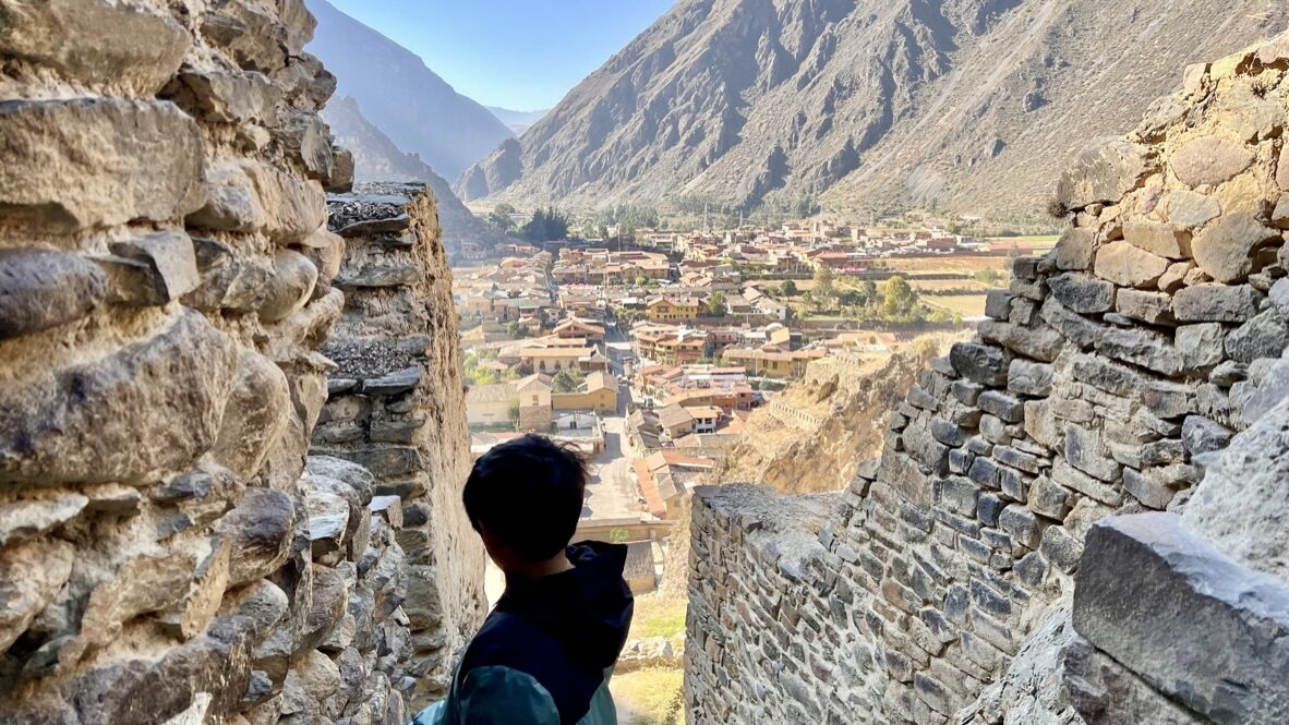 Boy looking between stone walls down at the town of Ollantaytambo in the valley