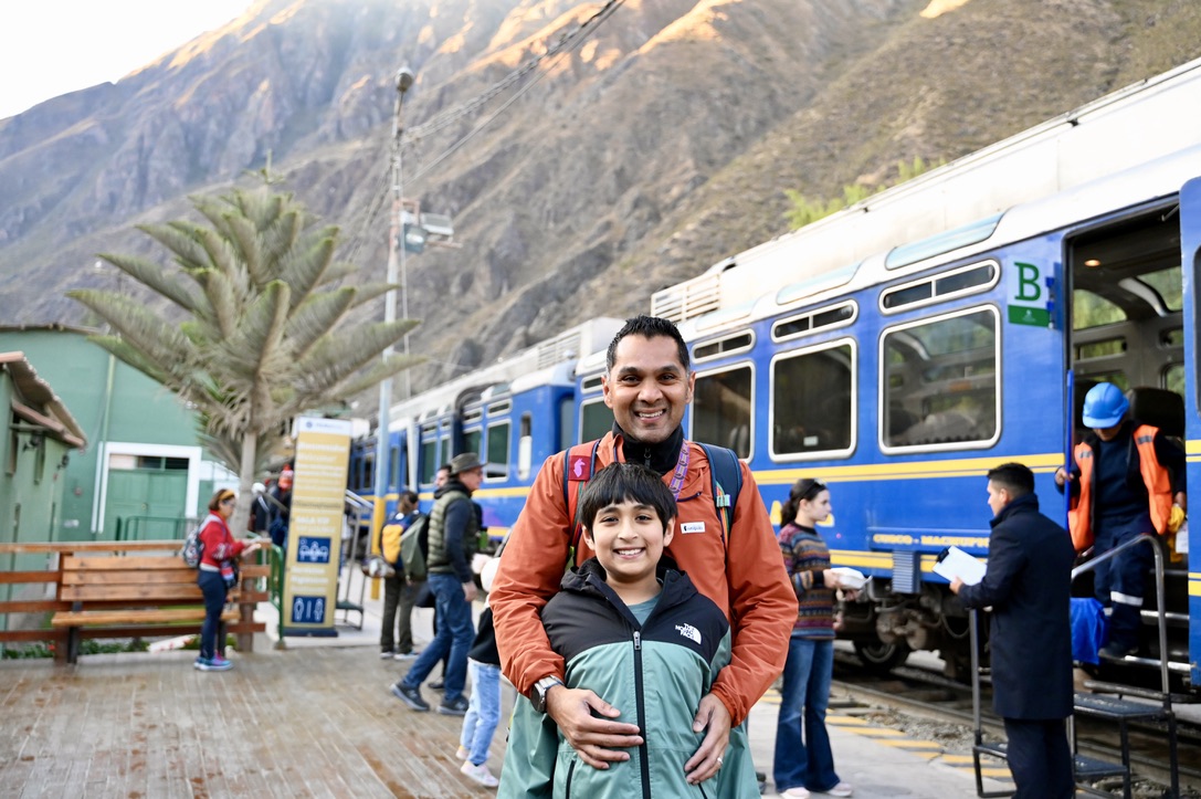 Man and boy smiling and standing on train platform, with a blue train with white roof and yellow stripes behind them and surrounded by mountains