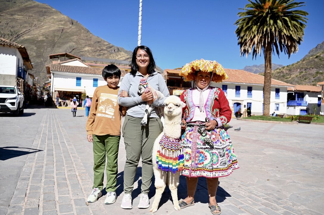 Woman and boy posing with a decorated alpaca and a woman in traditional dress in a town square