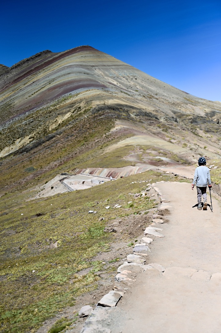 Boy with hiking pole walking down a mountain path with rainbow colored mountains with some level of wild grass and weed covering