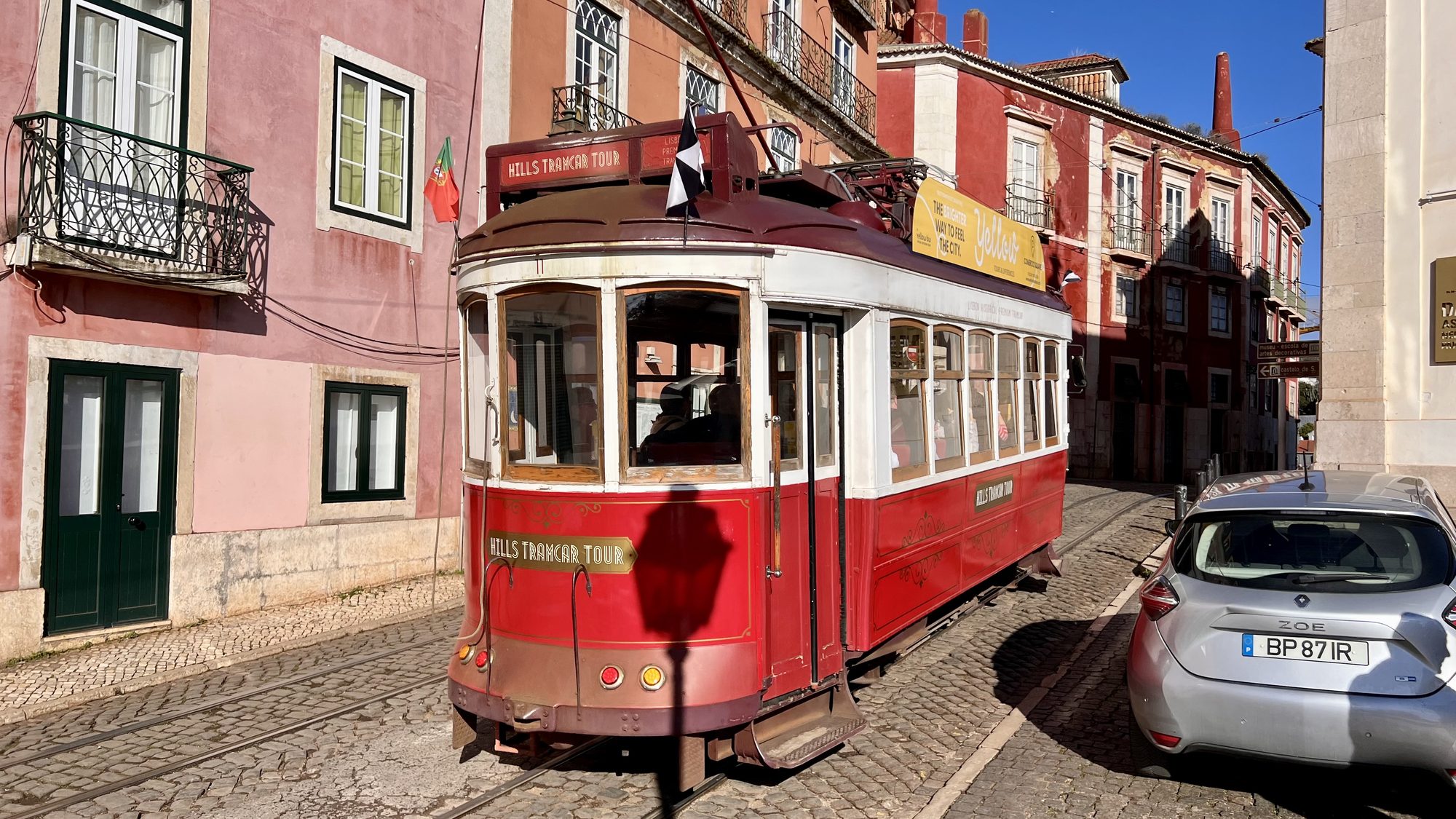 A red vintage tram rounding a corner on a cobbled street between pink and red buildings.