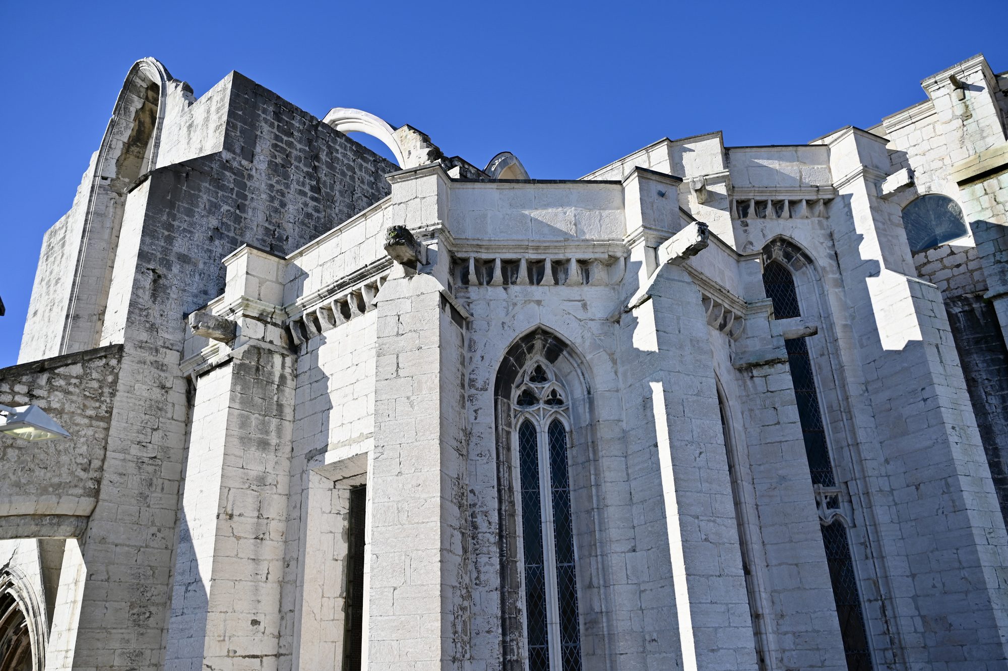 The pale stone walls, buttresses and arched windows of the Carmo Convent ruins against a bright blue sky.
