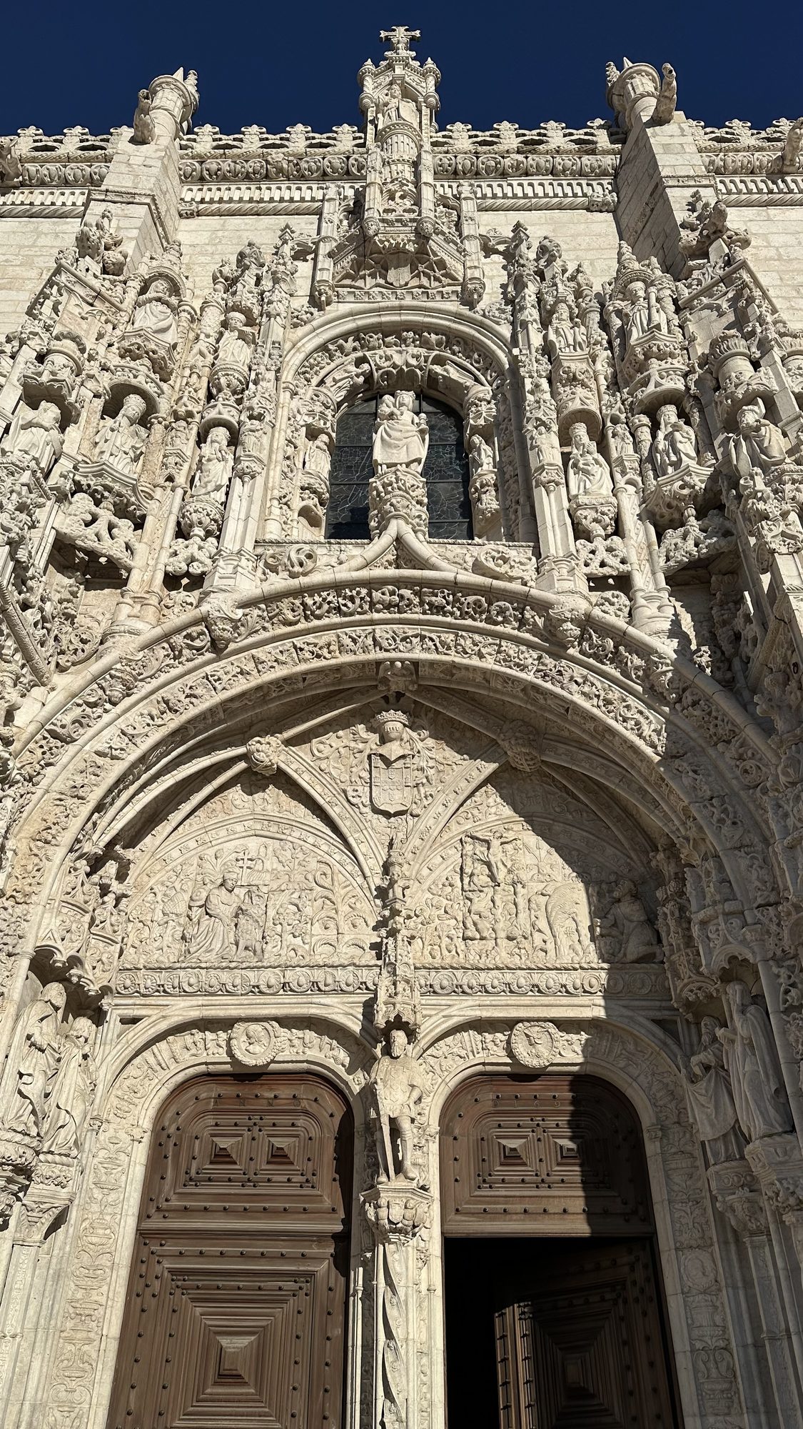 The ornately carved stone doorway of the Jerónimos Monastery, covered in statues and stonework, under a deep blue sky.