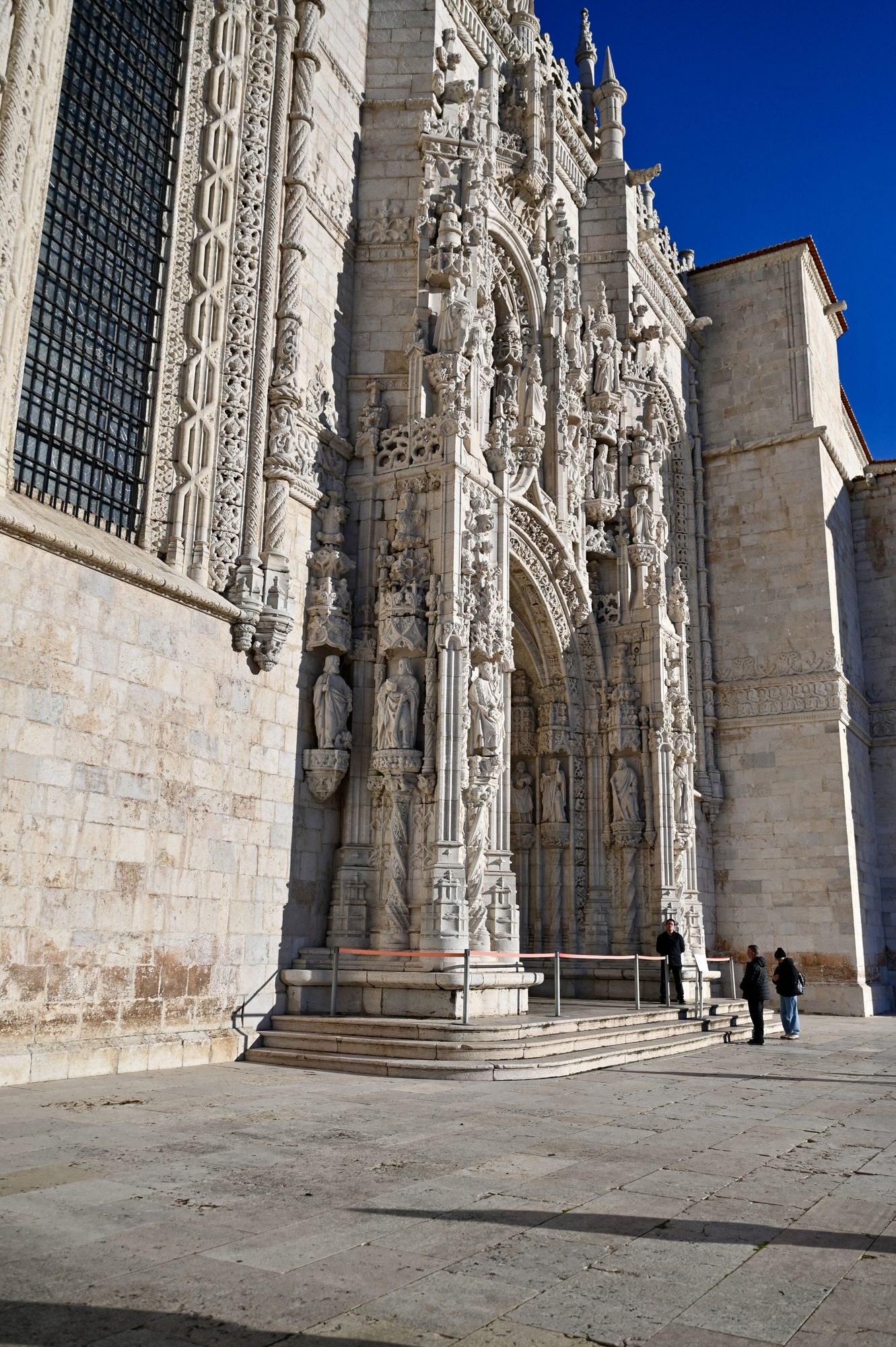 The richly carved white stone side doorway of the Jerónimos Monastery, under a deep blue sky.
