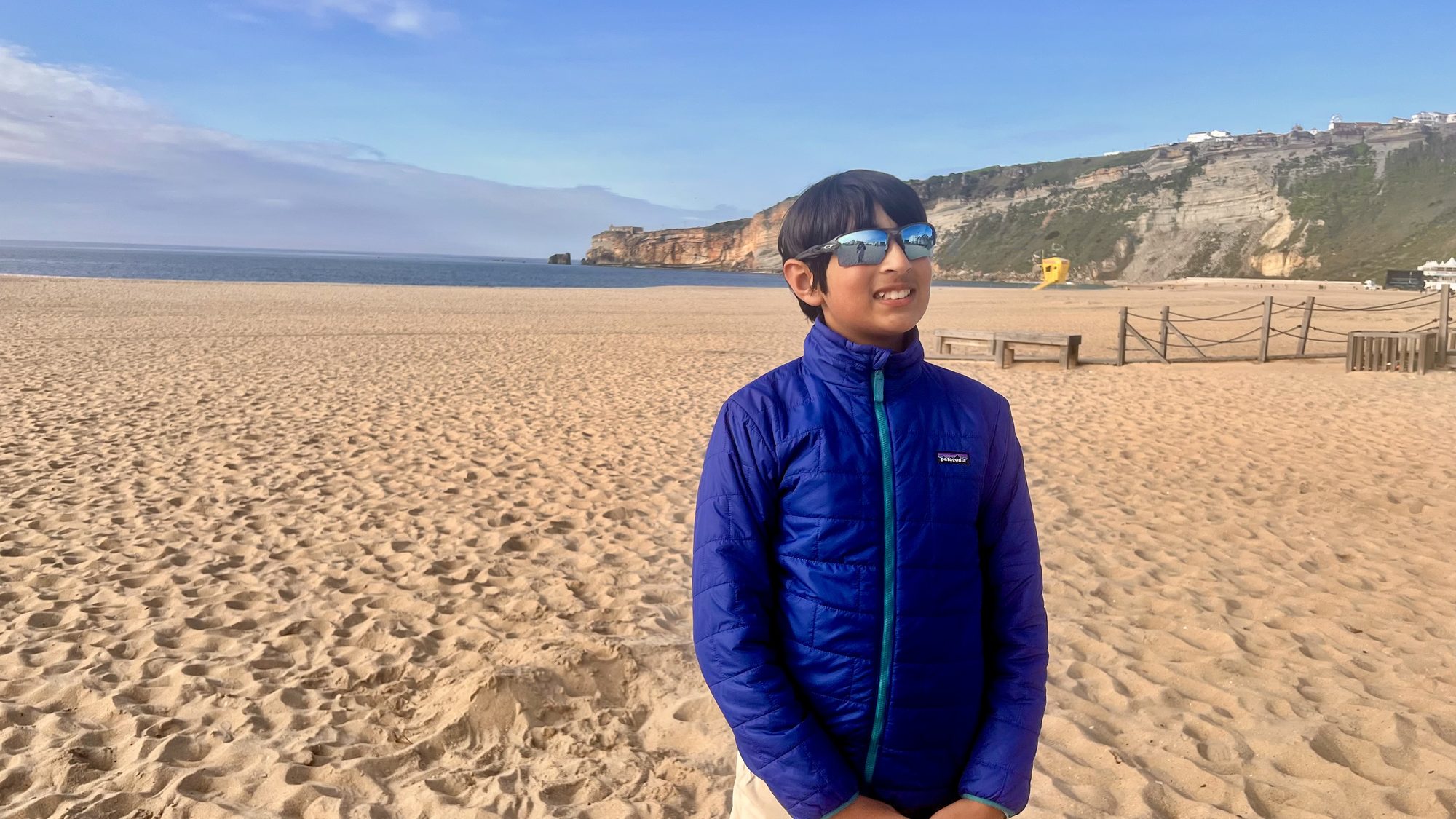 Our son in a blue jacket on the wide sandy beach at Nazaré, with the cliffs behind.