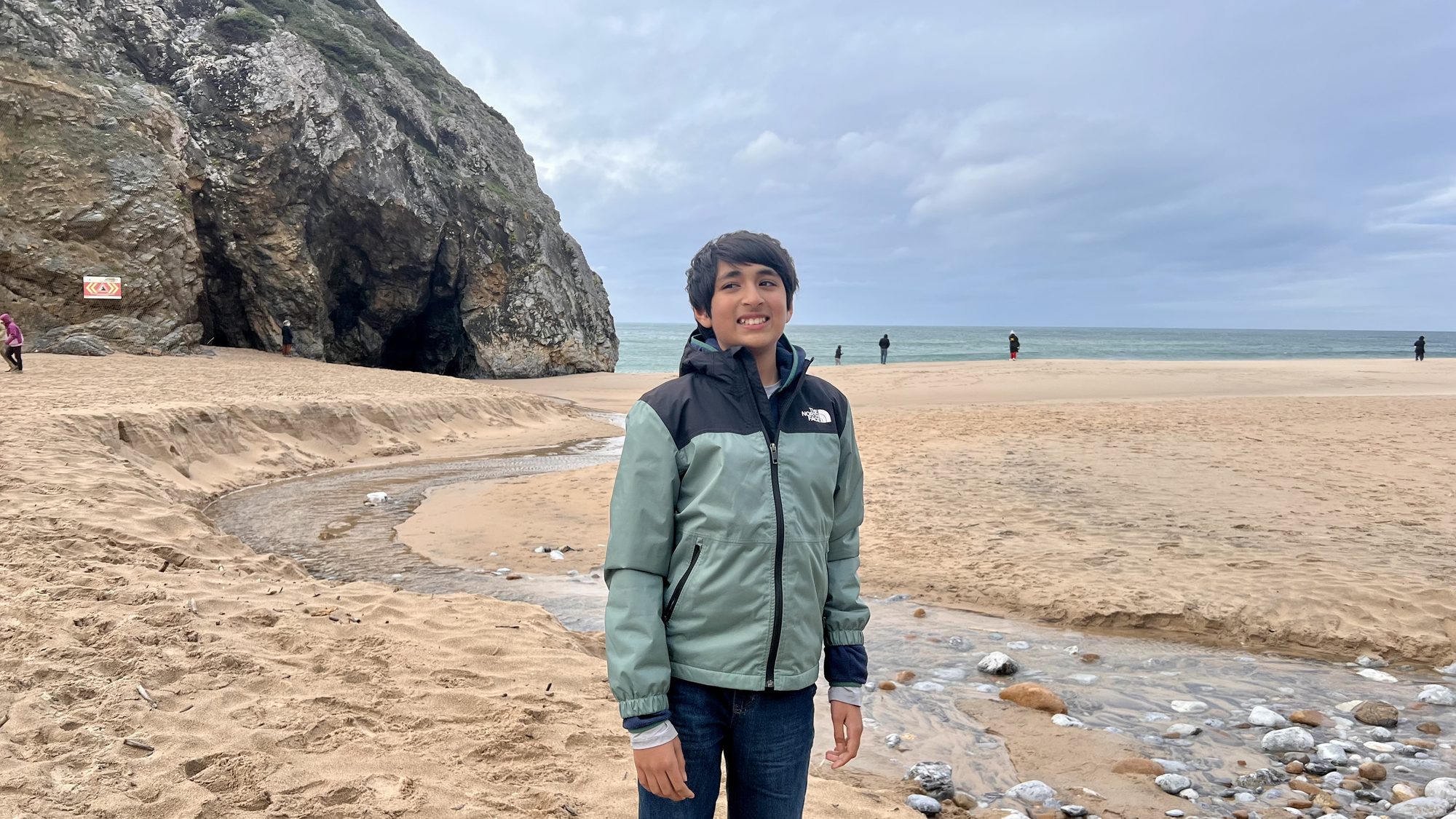 Our son standing by a small stream crossing the sand at Praia da Adraga, with cliffs behind.