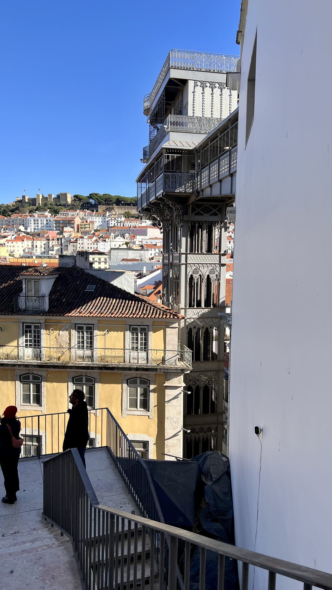 The iron tower of the Santa Justa Lift seen from a stairway, with the castle hill behind.