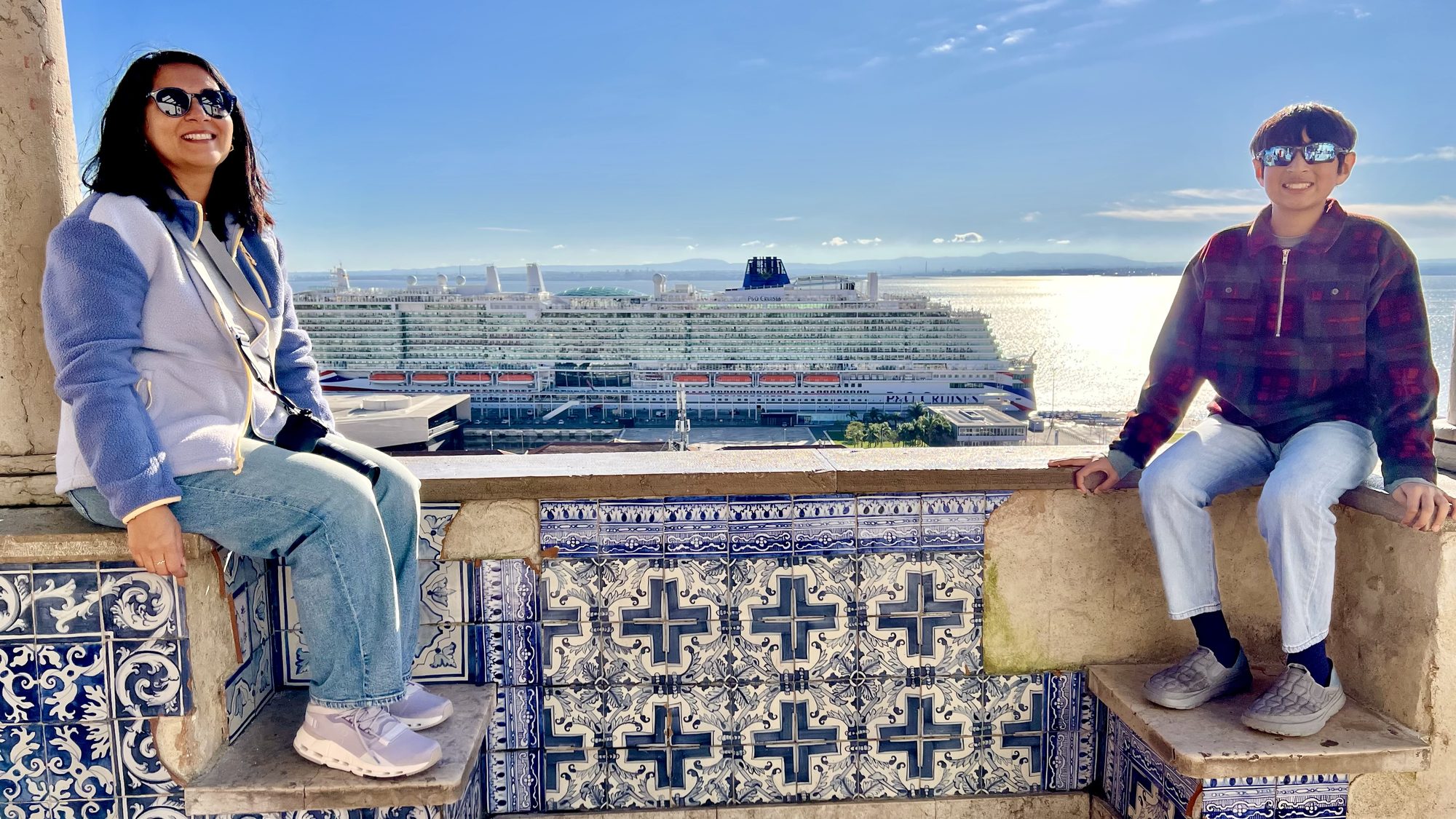 Two of us sitting on a blue-and-white tiled wall at Santa Luzia, with a cruise ship and the river behind.