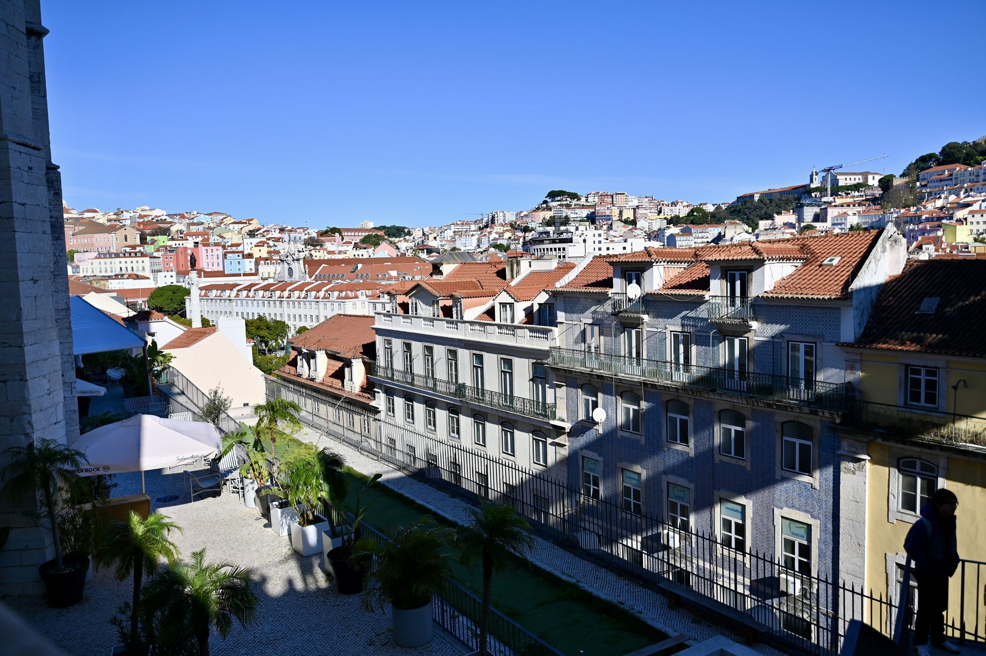 Lisbon’s rooftops and hills seen from a terrace with potted palms, near the top of the Santa Justa Lift.