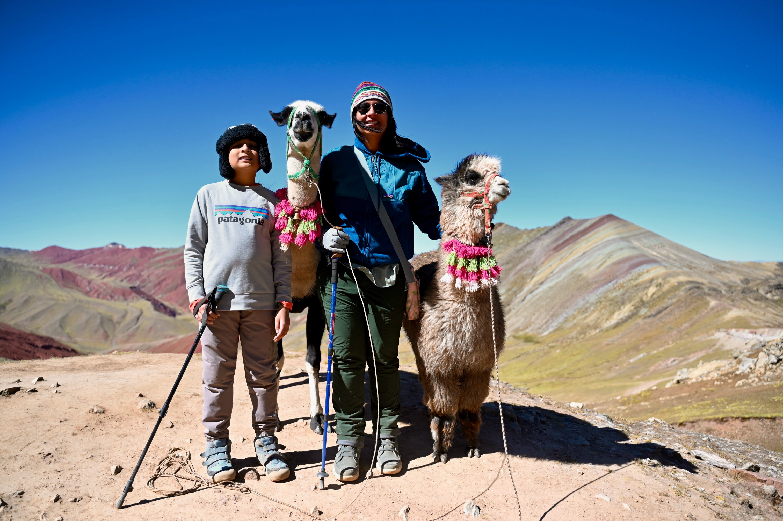 A parent and child on the trail at Rainbow Mountain in Peru, a llama standing beside them.
